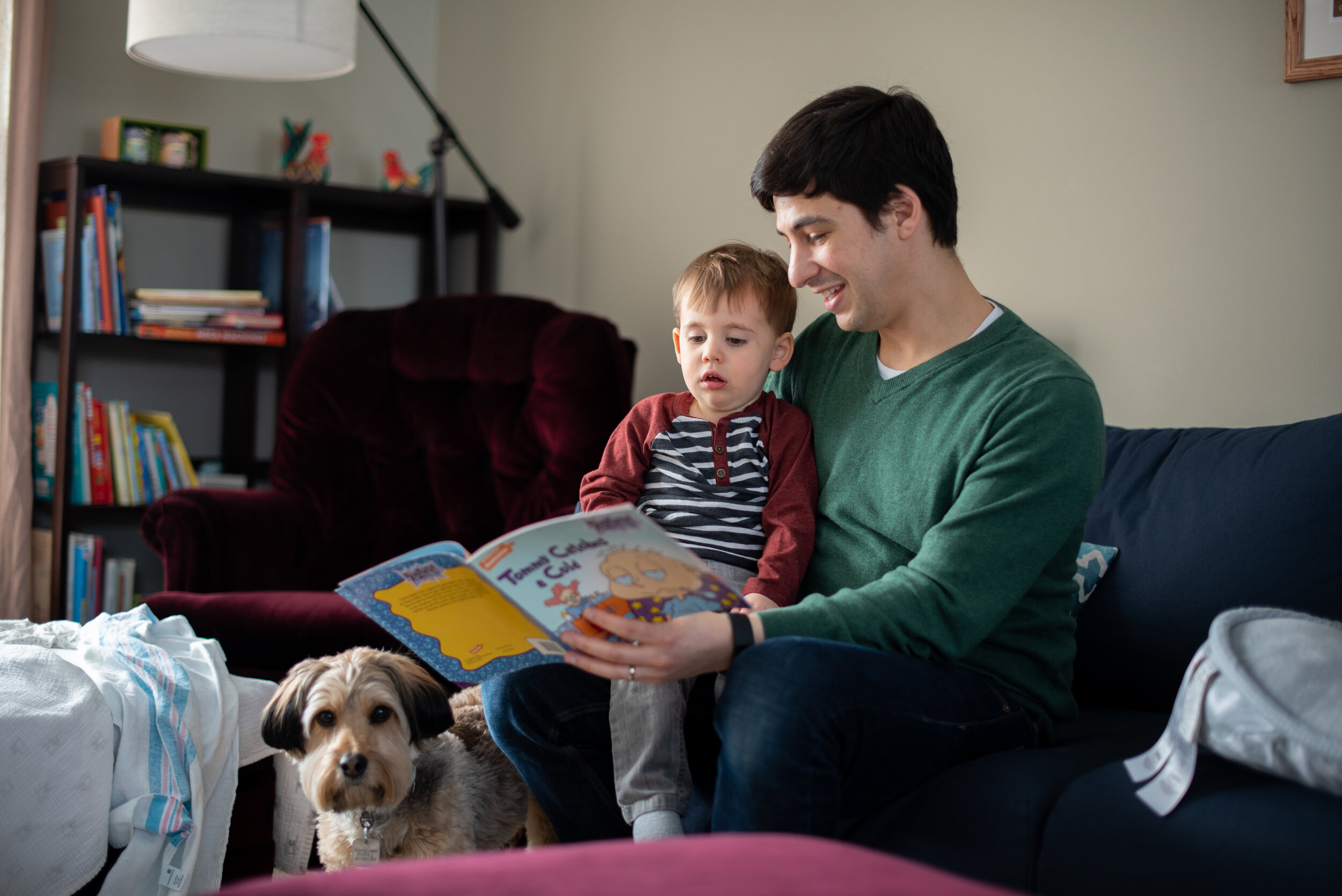 dad sitting on the couch next to a bookshelf with his son on his lap while reading a book to his son and dog during an in-home newborn lifestyle session