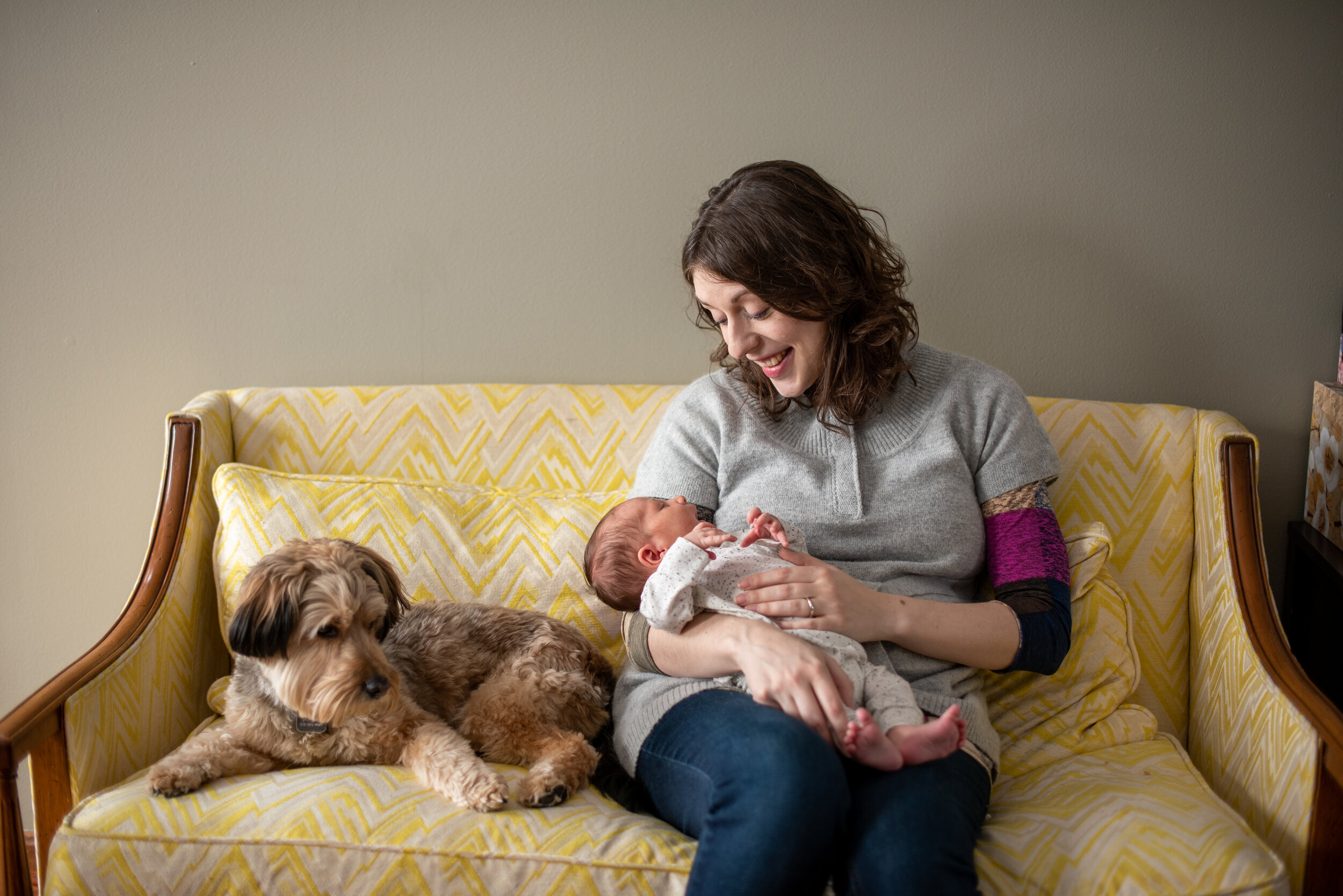 an in-home newborn lifestyle session in strongsville ohio of mom sitting on yellow and white striped couch next to her dog while holding her newborn baby and smiling down at him