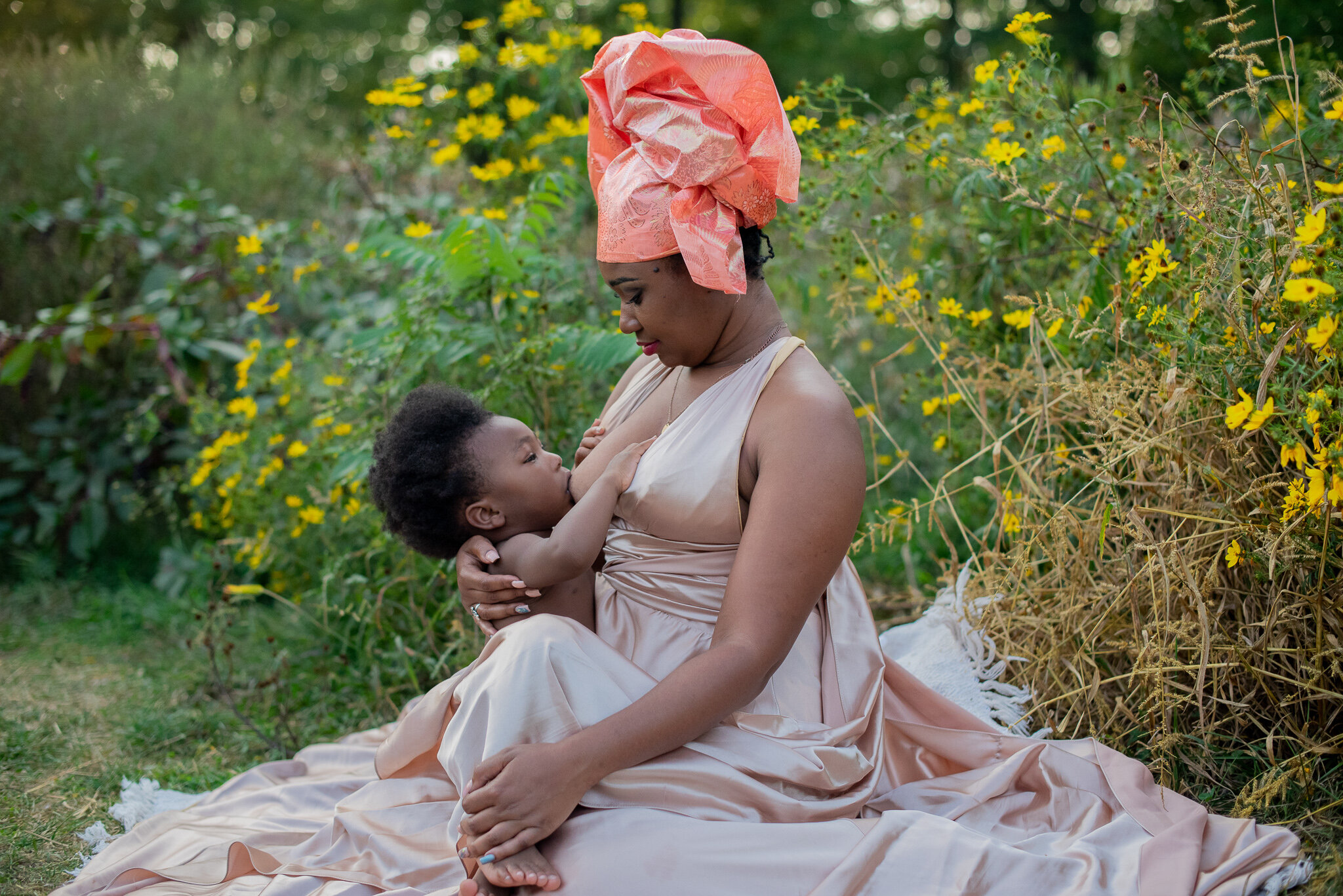 mother sitting in a field of yellow flowers while looking down at her breastfeeding child in Nigerian outfit