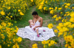 black mother in a white dress sitting in a field of yellow flowers breastfeeding her daughter in northeast ohio