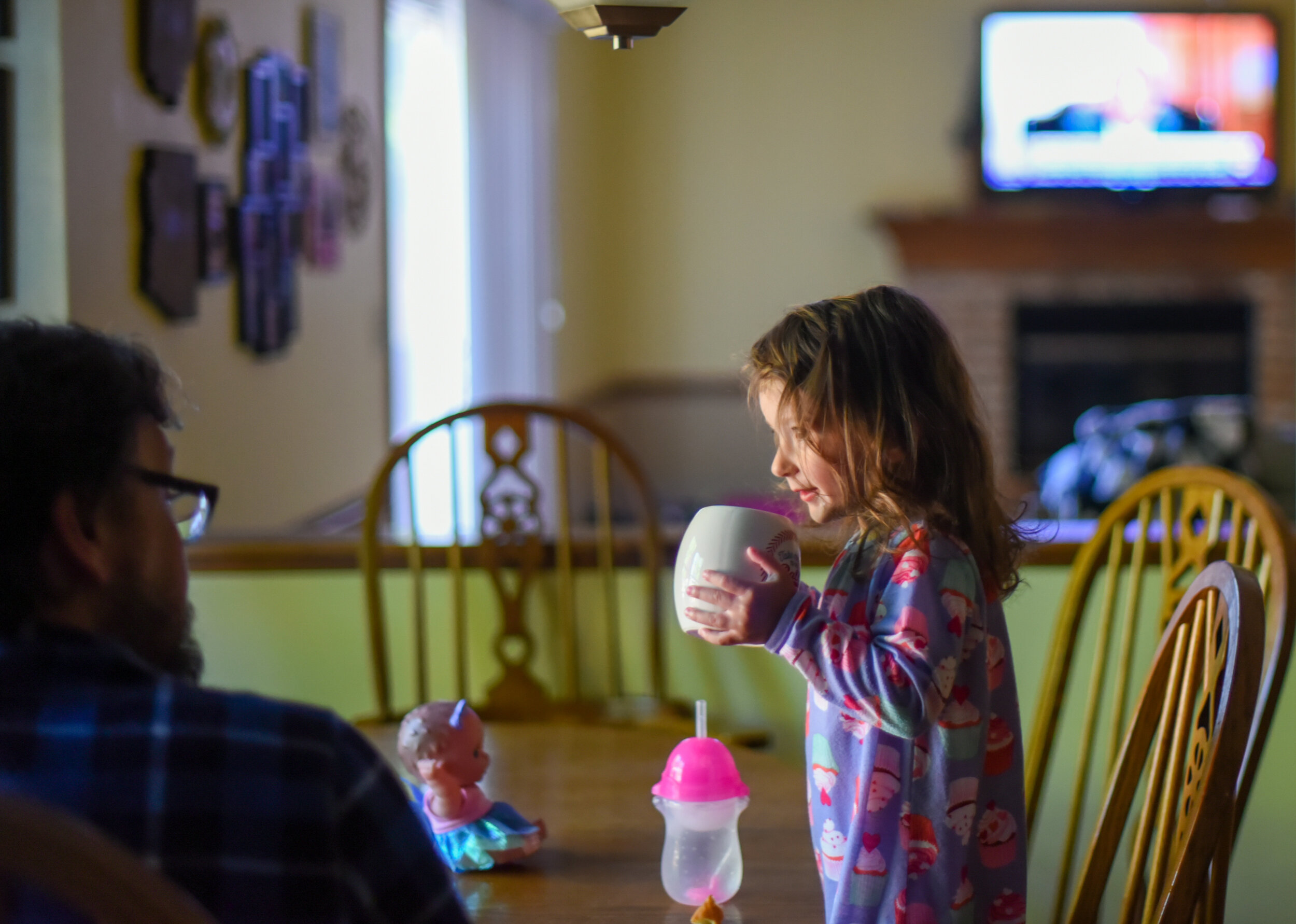 little girl sitting at the dining room table with her dad while drinking water out of a coffee mug and watching tv