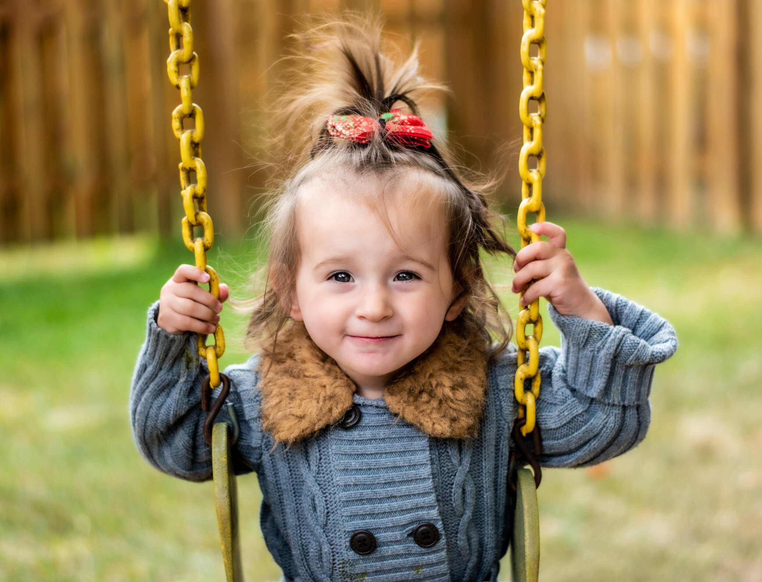 girl in a grey button up shacket with brown furry collar and a green and white plaid Christmas bow in her ponytail who is smiling at the camera while sitting on a yellow swing outside