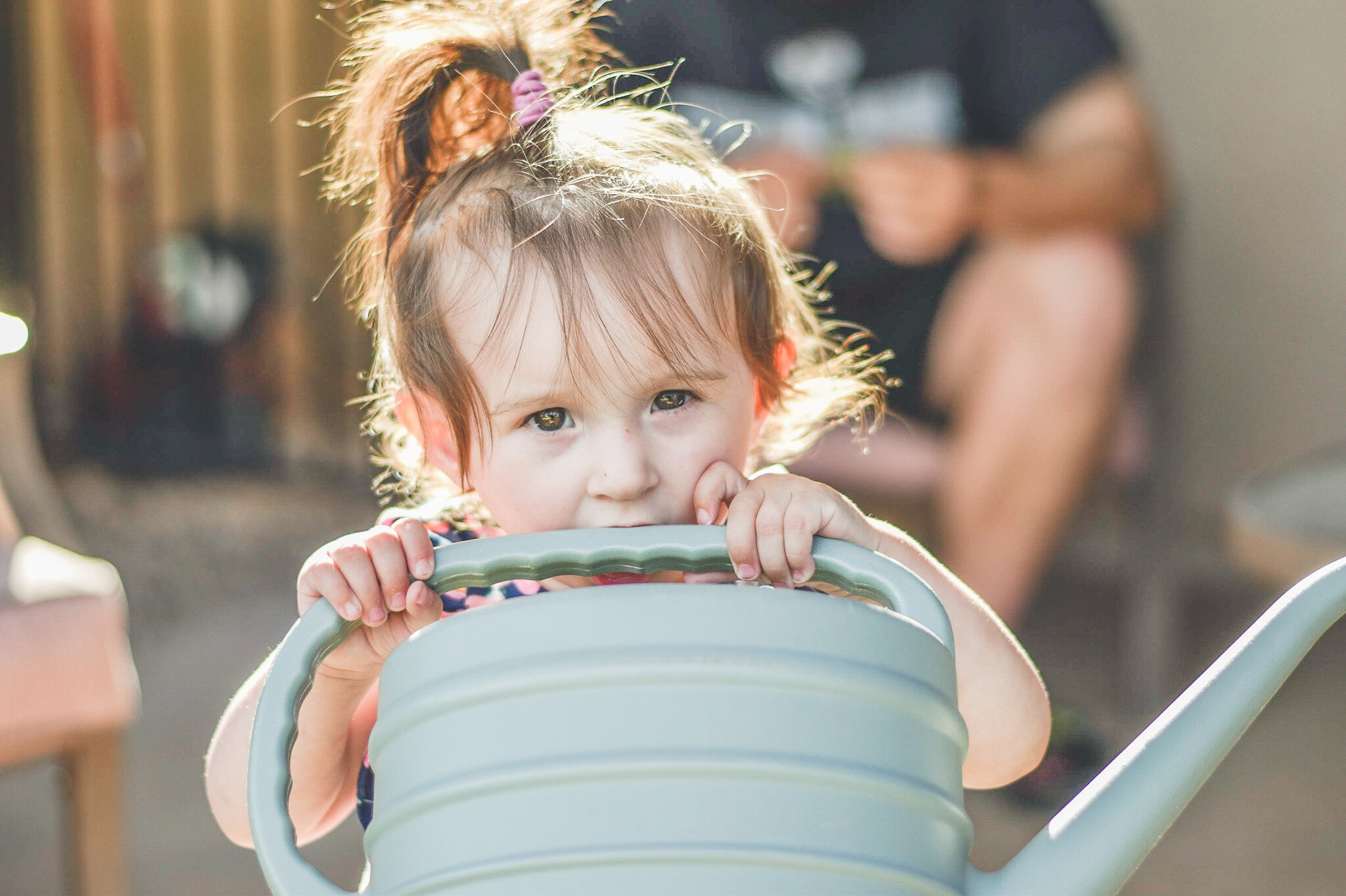 little girl with a messy ponytail staring at the camera while putting a green watering can up to her mouth