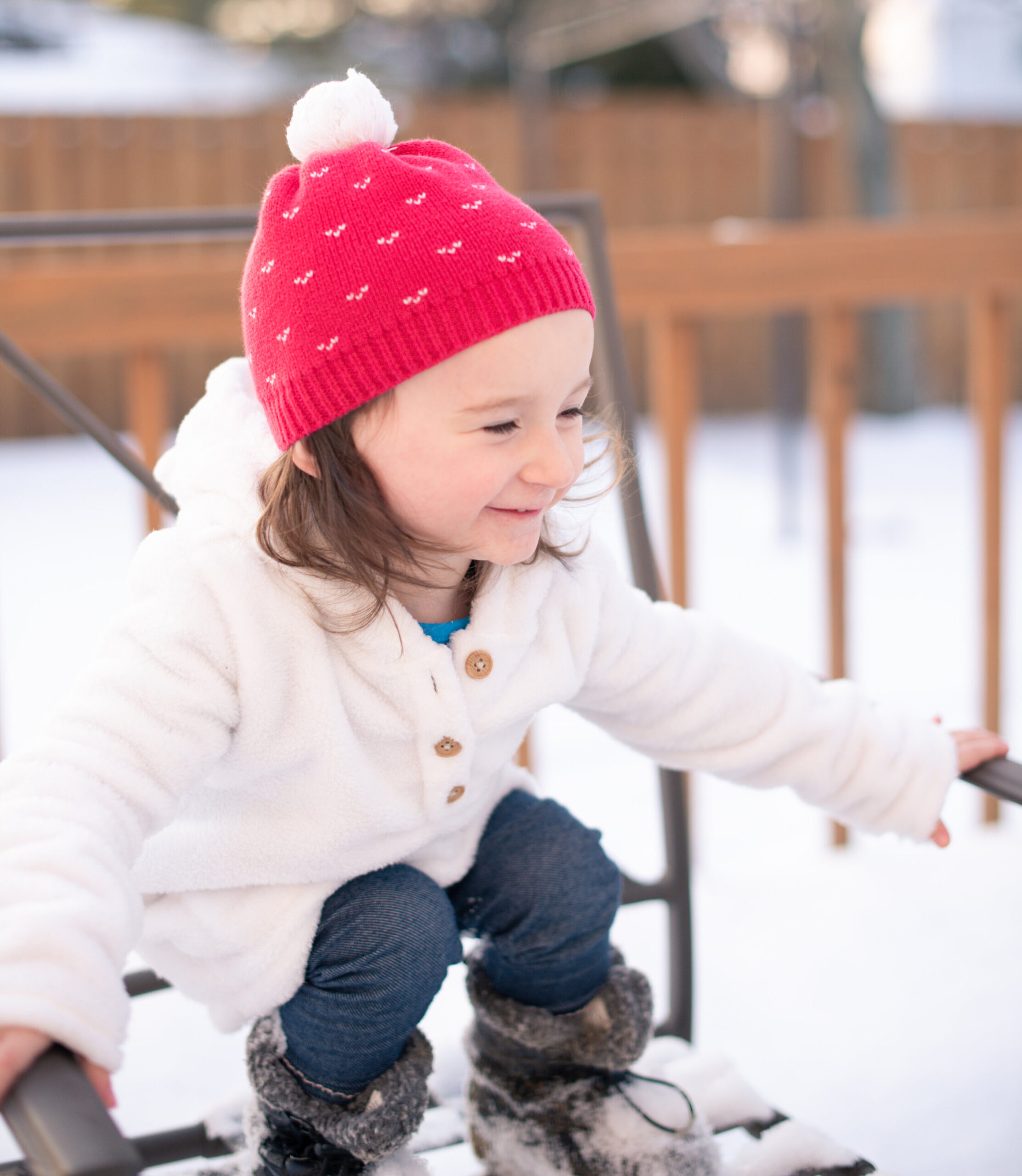 little girl with red and white winter hat, white button up sweater, blue jeans and black fuzzy boots standing on a snow-covered chair smiling 