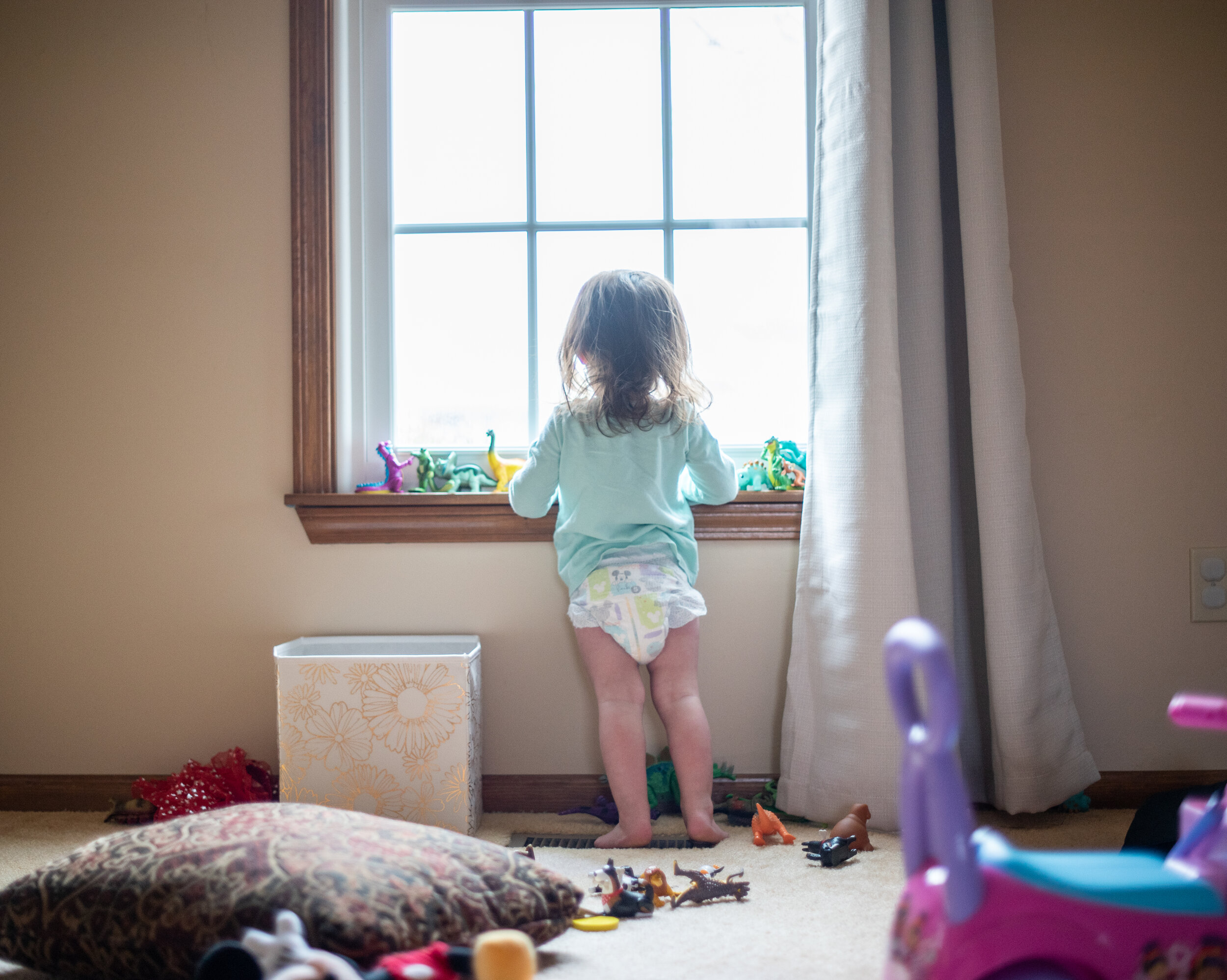 little girl in a blue shirt and diaper looking out the window with dinosaurs lined up along the windowsill