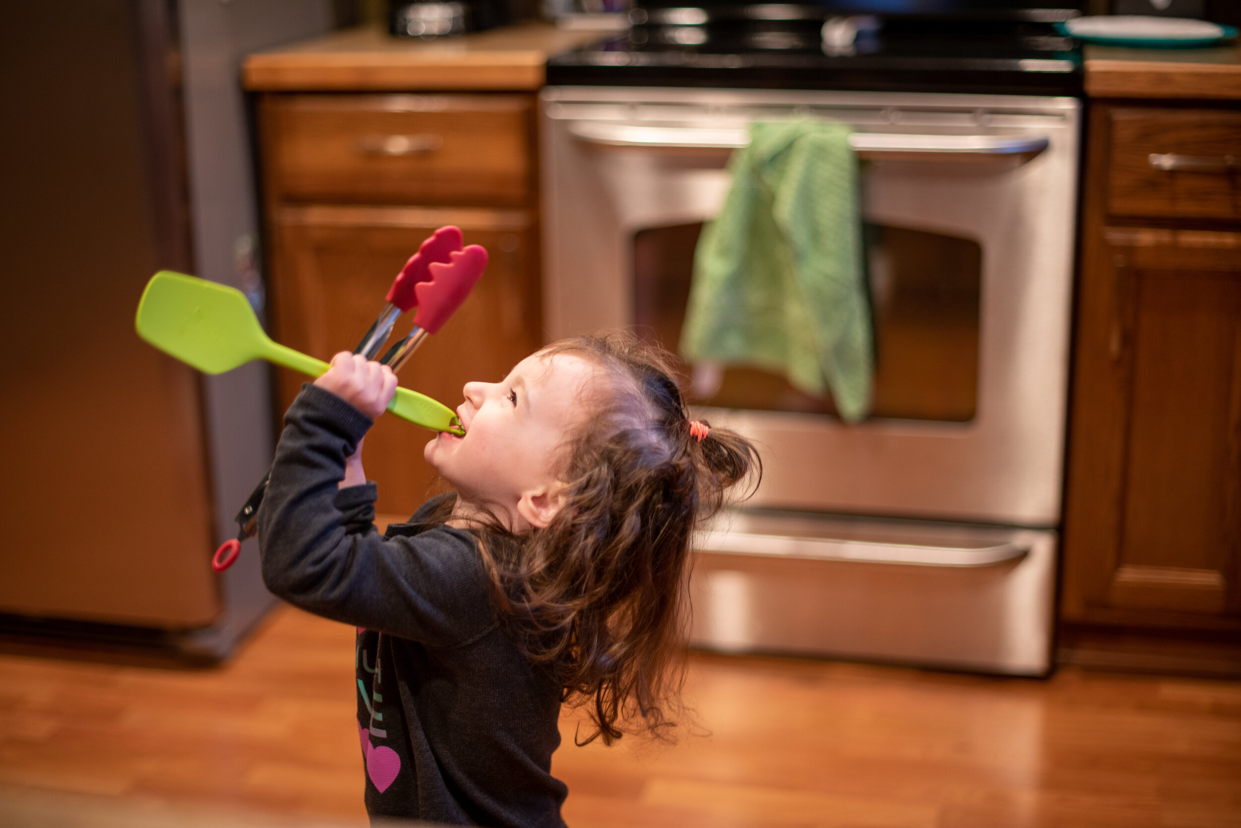 little girl in a kitchen holding red tongs and singing into a green spatula 
