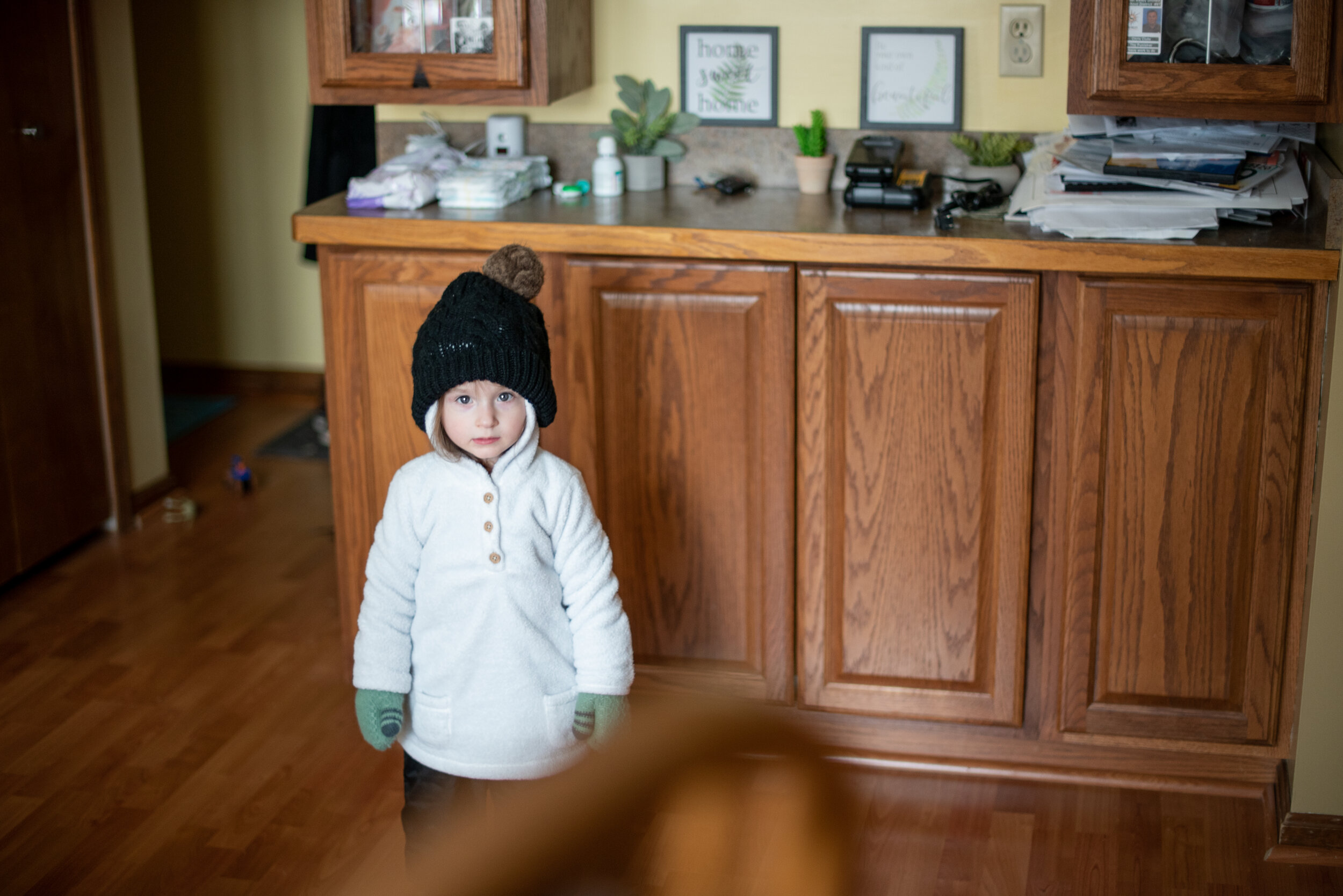 little girl in white sweater, black beanie, green mittens and black pants and looking at the camera without smiling
