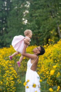 mom lifting little girl up as she laughs and they stand in a field of yellow flowers 
