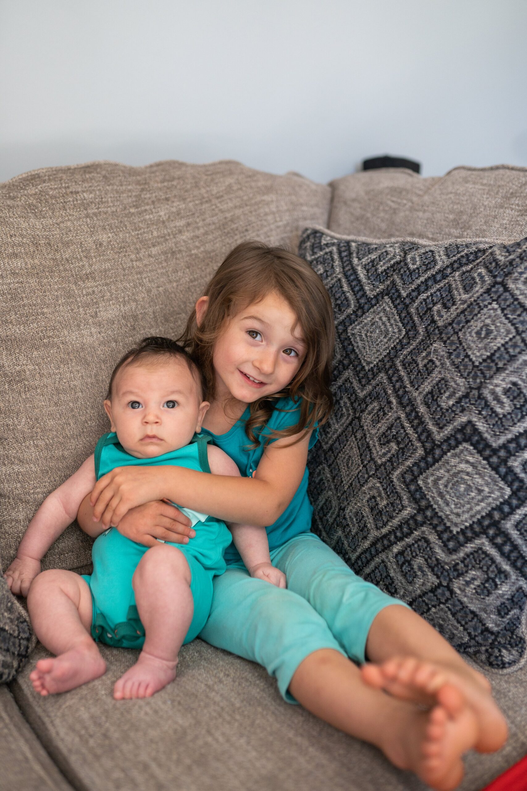 little girl sitting on a grey couch embracing her 3 month old little brother