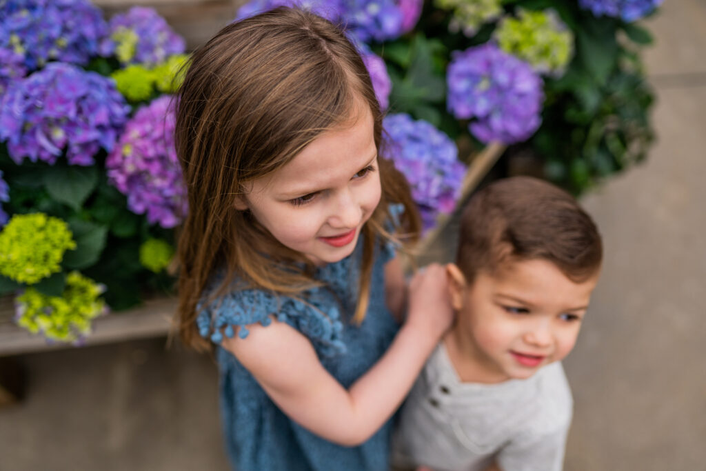 sister and brother staring off in a greenhouse filled with blue flowers 