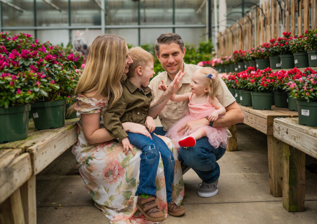 family in a greenhouse with mom and dad squatting down and dad has daughter on his lap, mom has son on her lap and they are trying to touch each others noses and laughing 