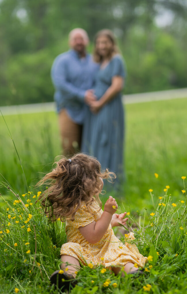 mom and dad standing in the background holding mothers pregnant belly and daughter tossing her head back to look at them with wild hair and sitting in the grass and yellow wildflowers 