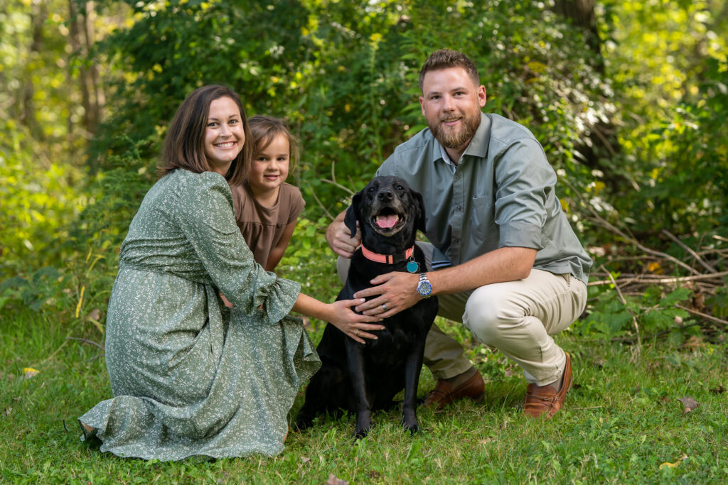 a smiling family with mom dad and daughter while petting their black family dog