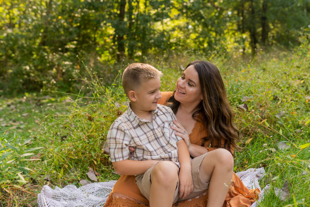 mom sitting in a grassy field laughing with her son sitting on her lap as her wavy hair done by a hair salon in cleveland ohio blows in the wind 