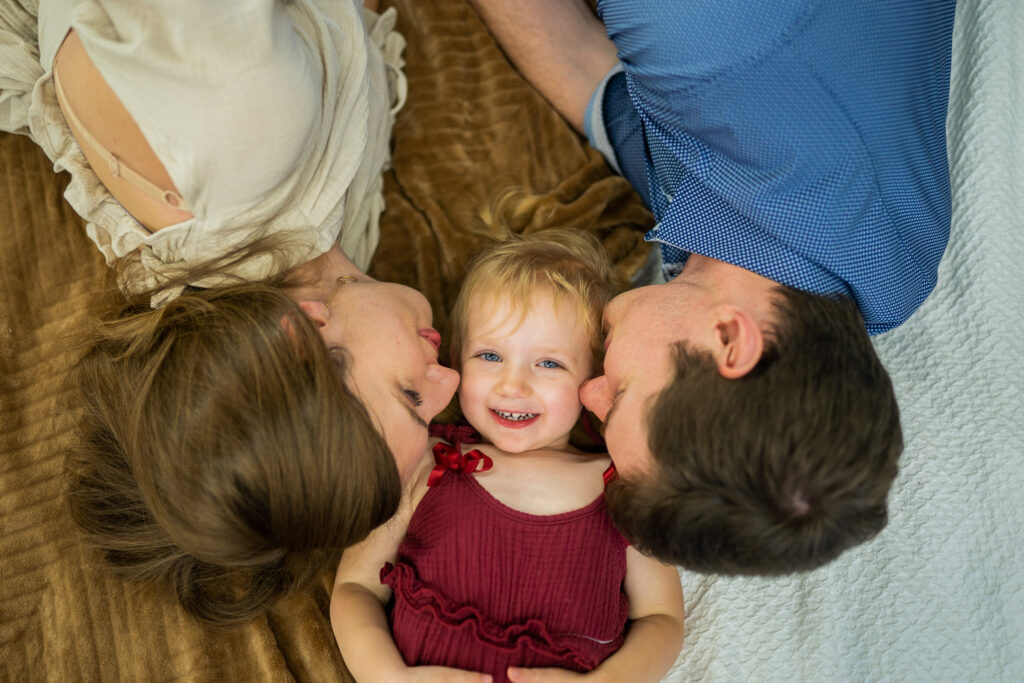 mom and dad laying down with daughter and kissing her on the cheek as she looks at the camera and smiles