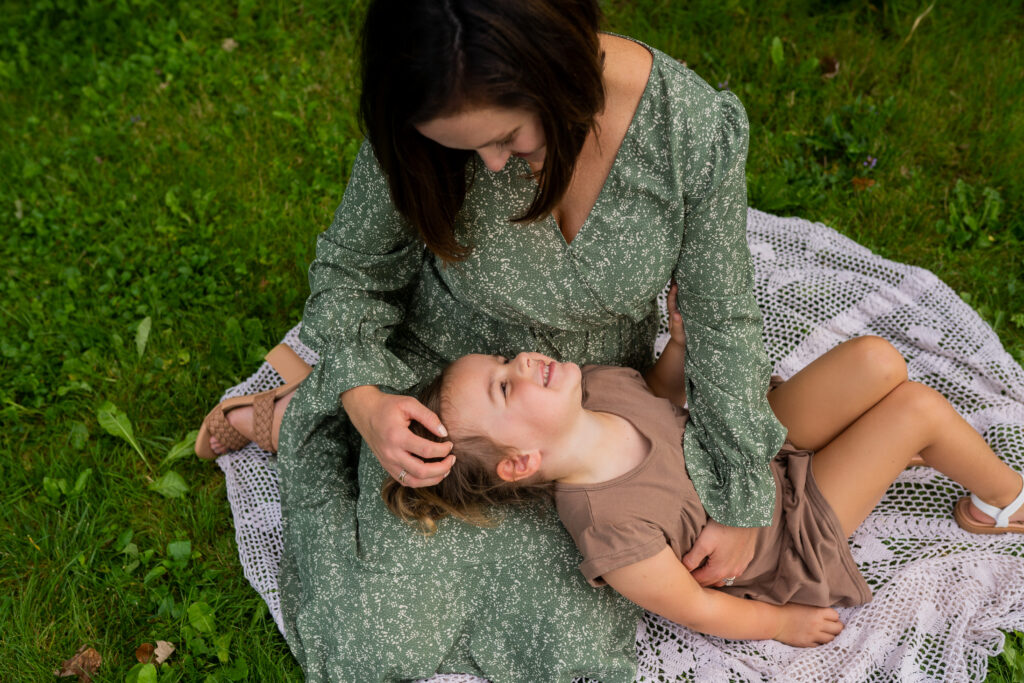 mom sitting in the grass on a crocheted blanket with her daughter laying down and her head on moms lap smiling at mom while moms hand runs through her hair 