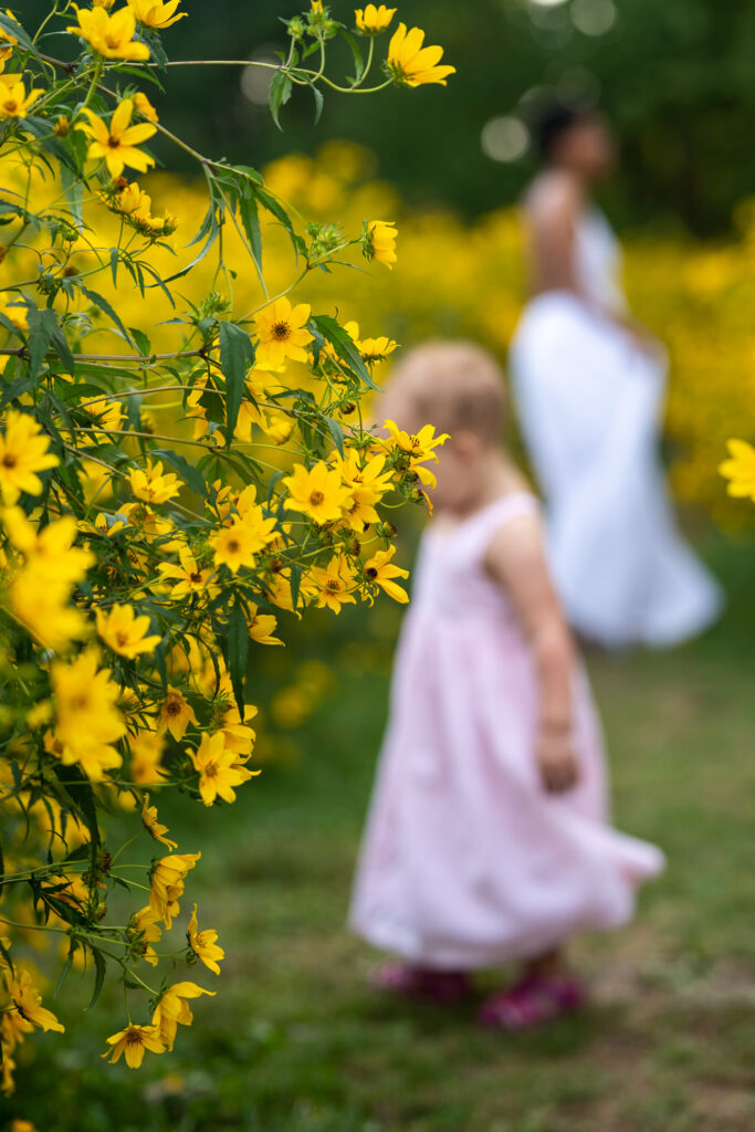 a field of yellow flowers with a mother and daughter dancing in dresses in the background