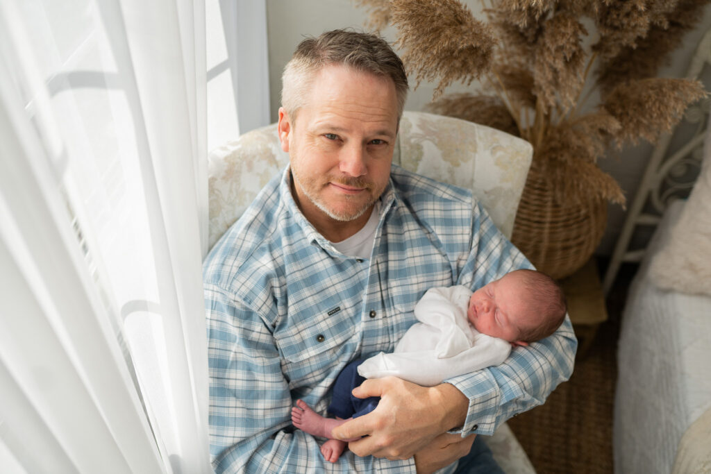 dad sitting in a white chair while holding newborn baby boy and looking up at the camera smiling