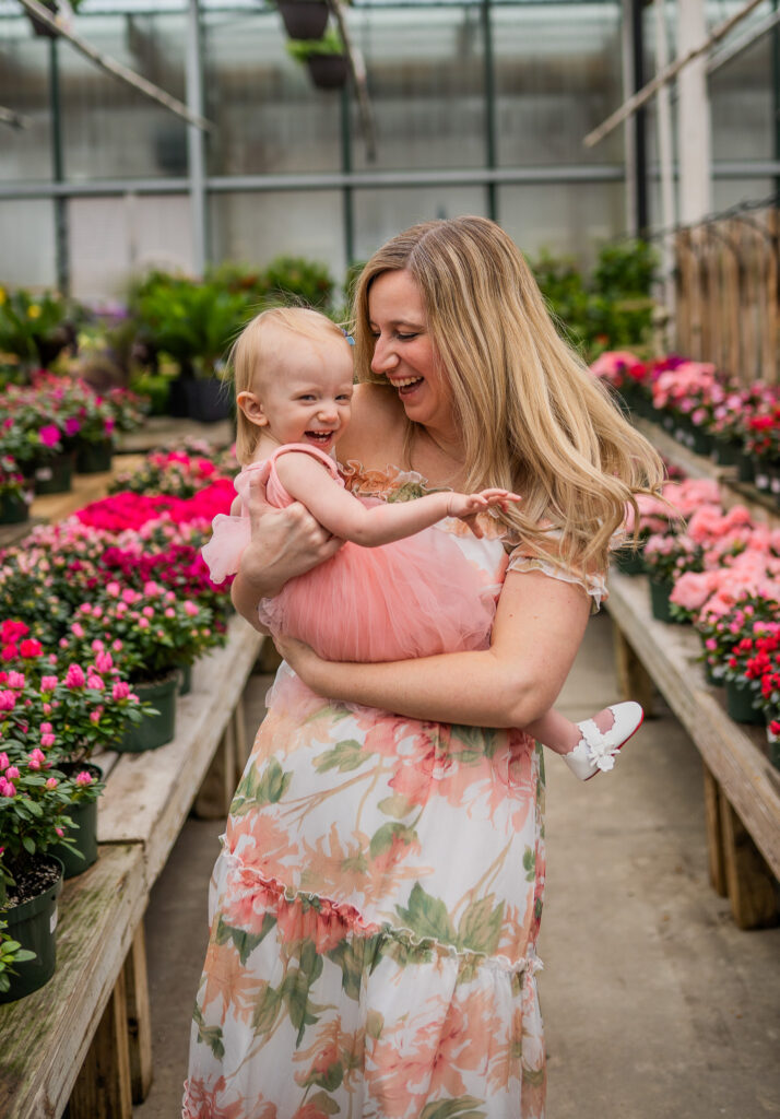 mom holding a one year old girl in a greenhouse and spinning around while mom and daughter laugh together as they spin