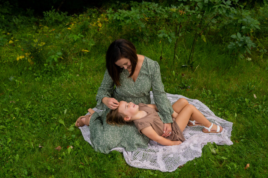 mom sitting on a blanket in the grass while her daughter lays her head on moms lap and mom runs her hands through her daughters hair as they smile at each other 