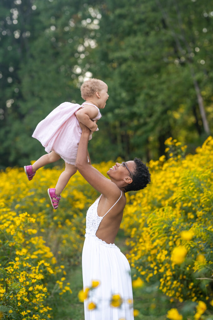 mom in a backless white dress holding up her daughter in a light pink dress as they smile at eachother in a yellowflower field in ohio