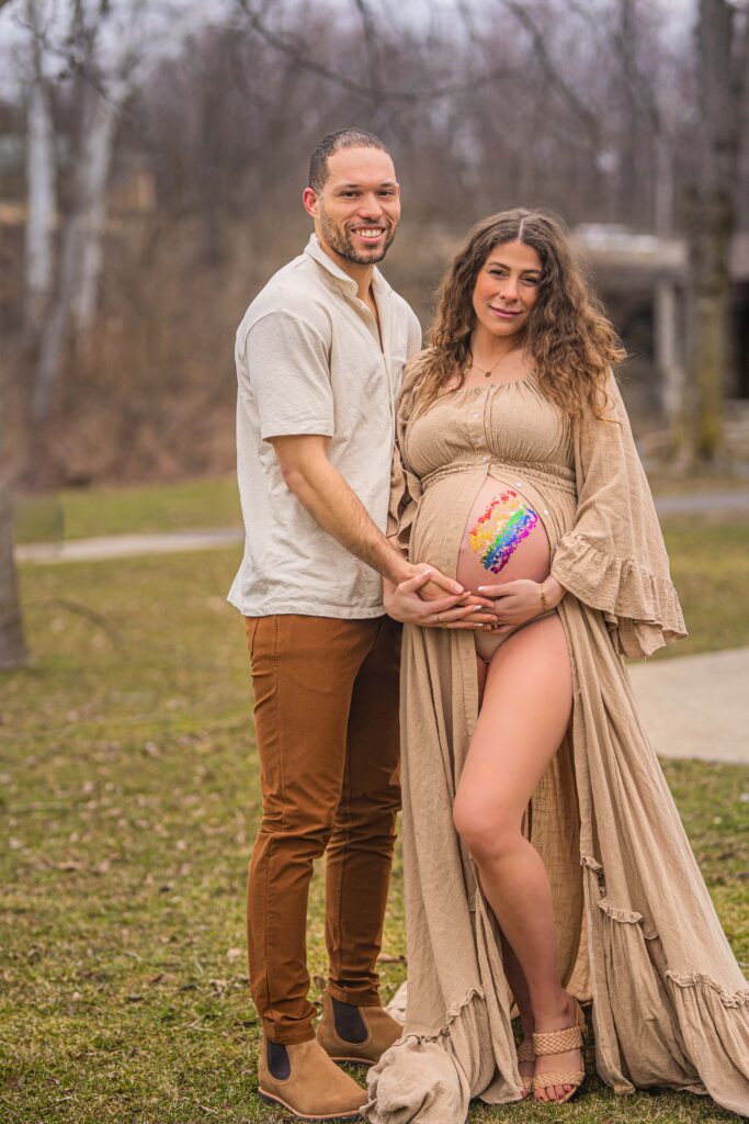 expecting mother and father smiling at the camera with a rainbow made of foil flecks are on her belly to represent a rainbow baby