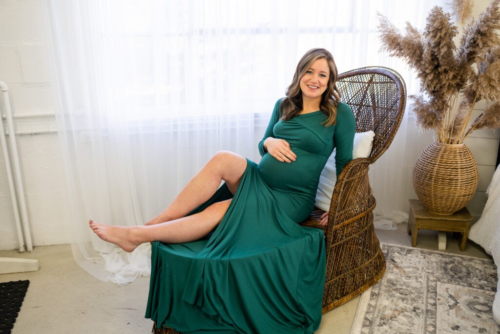pregnant mom in emerald green dress sitting on a rattan chair with her hand on her baby bump while smiling at the camera 