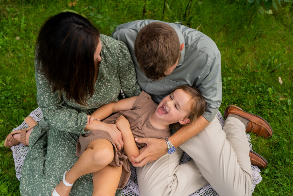 mother and father sitting in the grass and tickling their daughter 