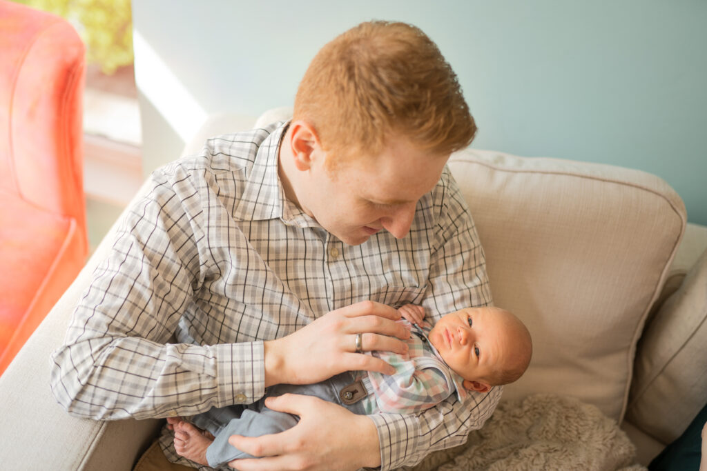 dad sitting on a grey cough in a plaid shirt while holding his newborn son and smiling down at him