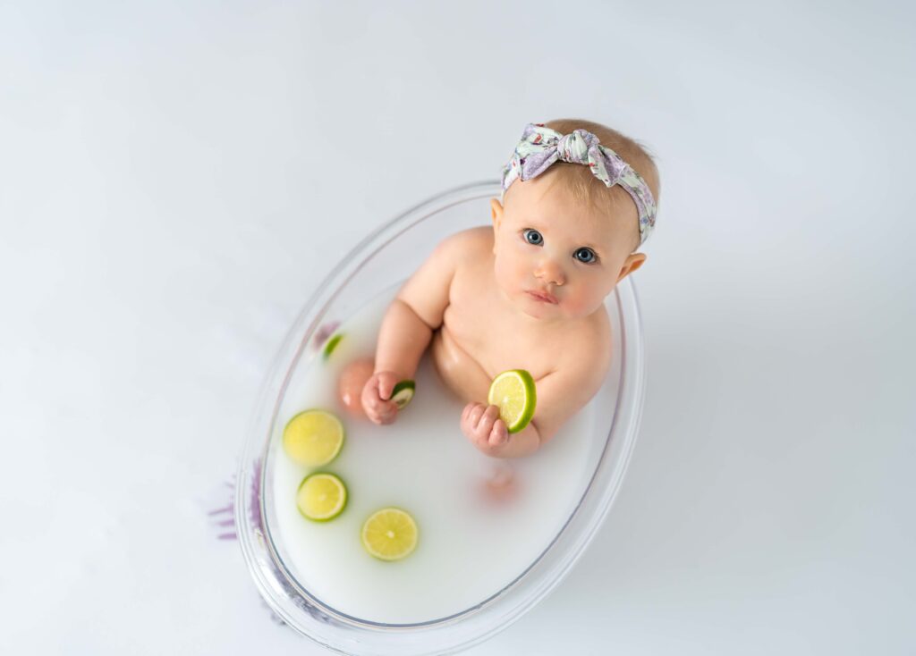 baby looking up at camera with a lime slice in her hand while sitting in a clear tub milk bath and lime slices