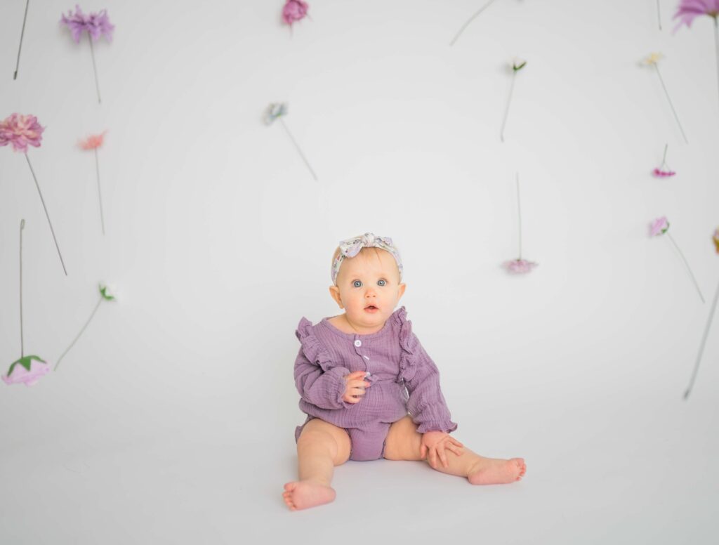 a baby in a lilac onsie and floating flowers around her staring curiously at the camera 