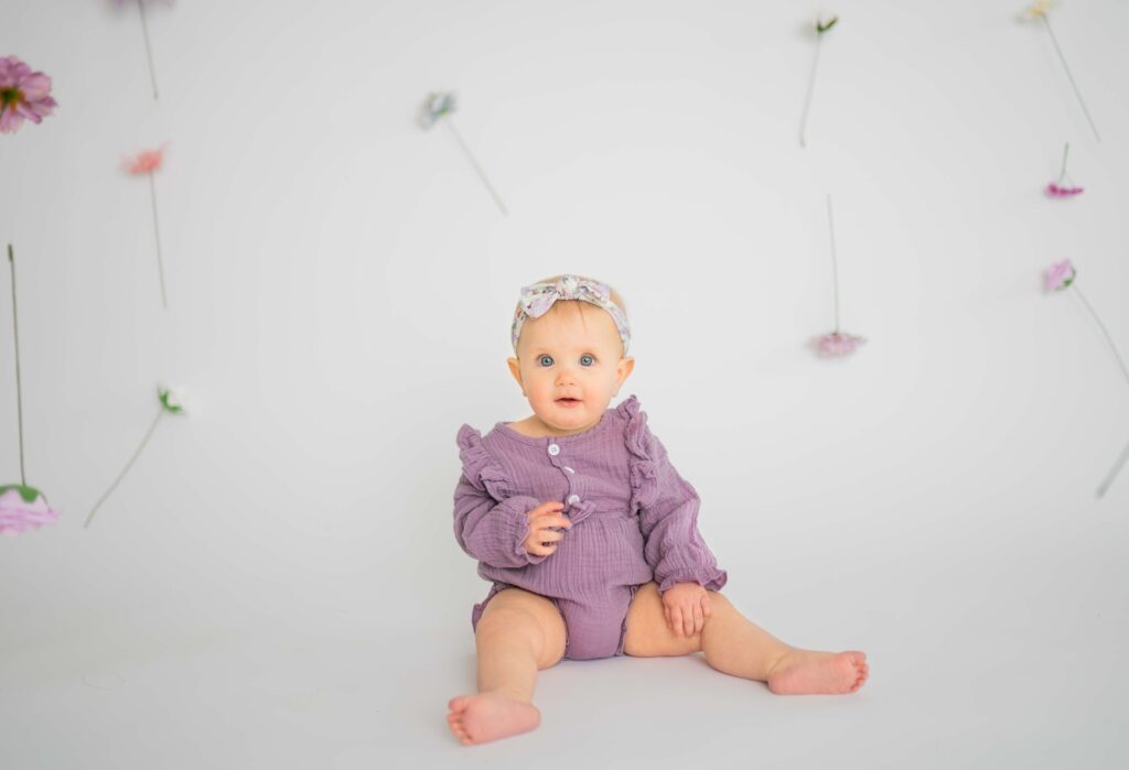 sitting baby girl in lilac onsie surrounding by floating flowers looking at the camera with her blue eyes and smiling 