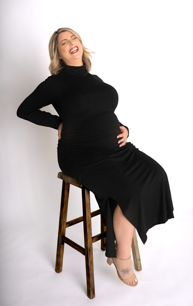 pregnant mother in fitted all black dress and white backdrop holding her belly and laughing