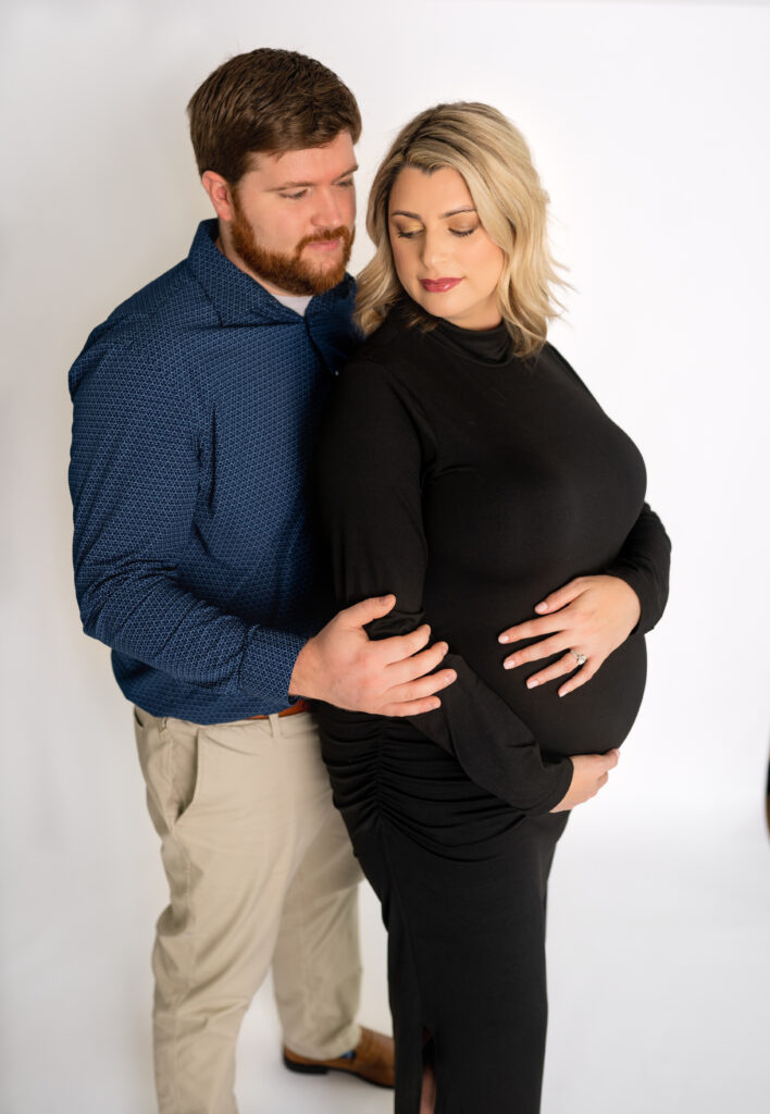 pregnant mother in fitted all black dress holding her belly with her husband in a blue shirt and khakis stands behind her holding her and gazing at her