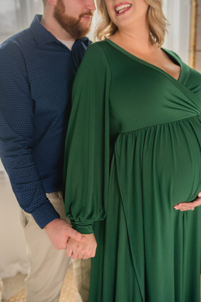 pregnant mother in green maternity dress with one hand holding her belly and the other holding her husbands hand as he stands behind her gazing at her in a blue shirt and khaki pants in medina ohio 