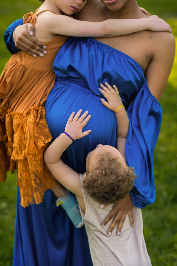 pregnant mother in blue dress holding her daughter as her toddler reaches up for her and puts hands on her belly