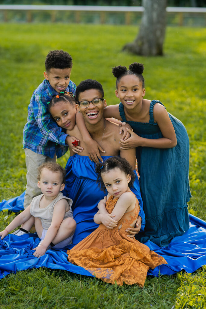 pregnant mother in blue dress sitting while being surrounding and hugged by all of her children as they laugh and smile together