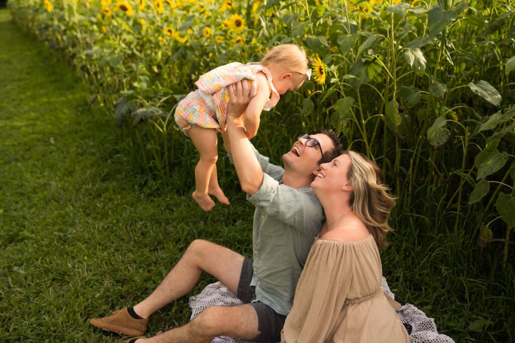 mom and dad sitting on a sunflower field holding their toddler up and smiling at her 